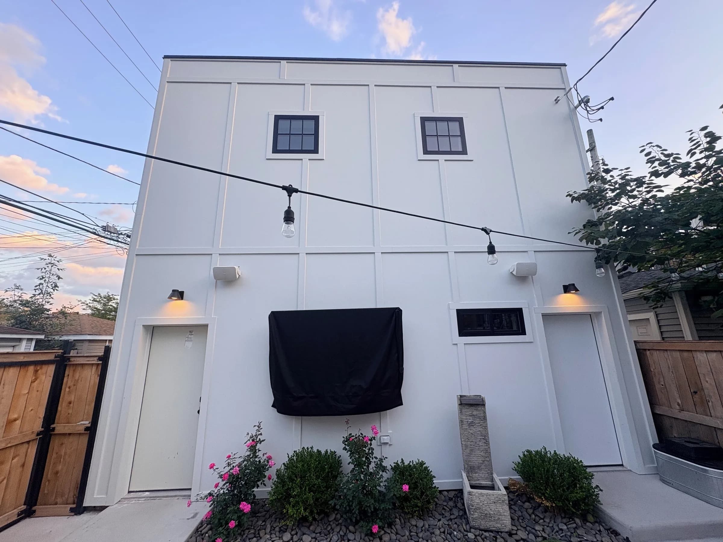Coach house at dusk: white board-and-batten siding, private entrances, covered grill, rose bushes and cafe string lights