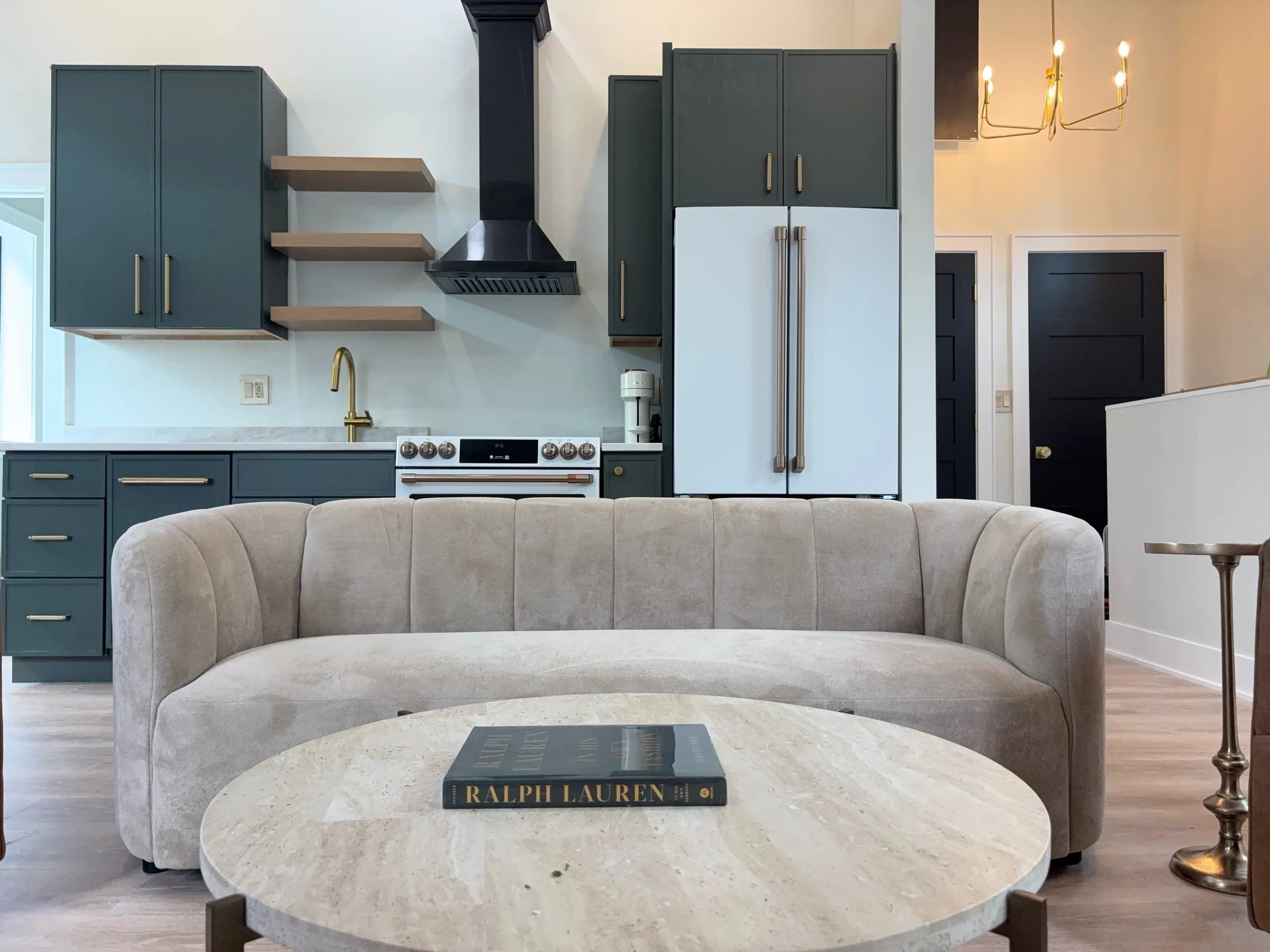 Channel-tufted sofa and round travertine coffee table facing the kitchen, with a brass chandelier overhead