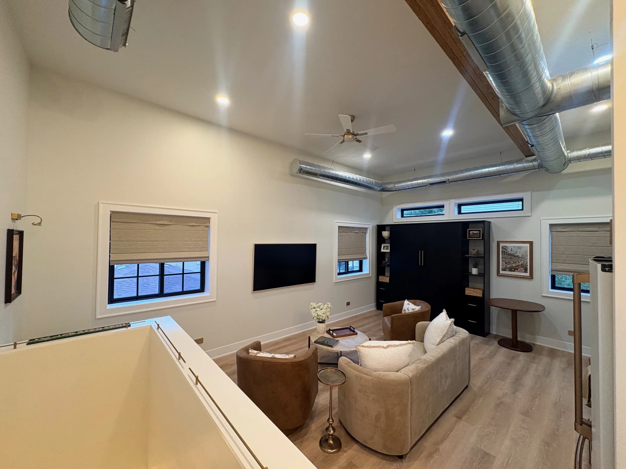 Living room seen from the stair landing, showing the vaulted ceiling, exposed spiral ductwork, woven shades and Murphy bed cabinet