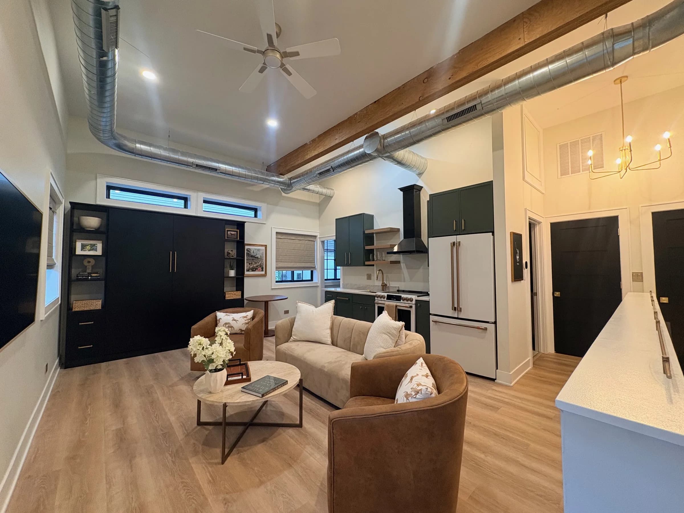 Wide view of the living room and kitchen: sofa and leather swivel chairs on a travertine coffee table, the black Murphy bed cabinet, green kitchen cabinets and a brass chandelier under exposed ductwork